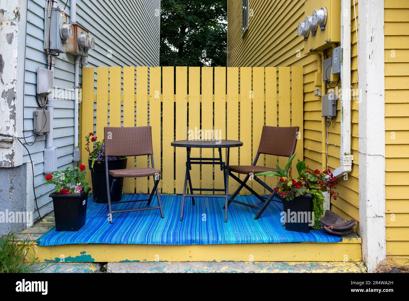 Due piccole sedie da patio e un tavolo rotondo tra due case con una recinzione gialla, fiori e un tappetino blu. Foto Stock
