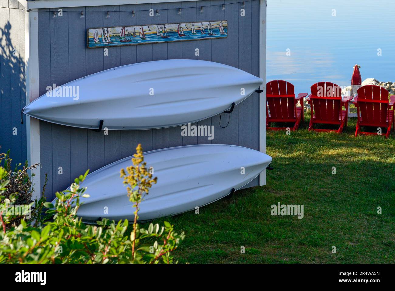 Due kayak bianchi appesi su un piccolo capannone di barche di colore grigio. C'è un cartello sopra l'edificio. Tre sedie rosse sono sull'erba e guardano verso l'oceano. Foto Stock