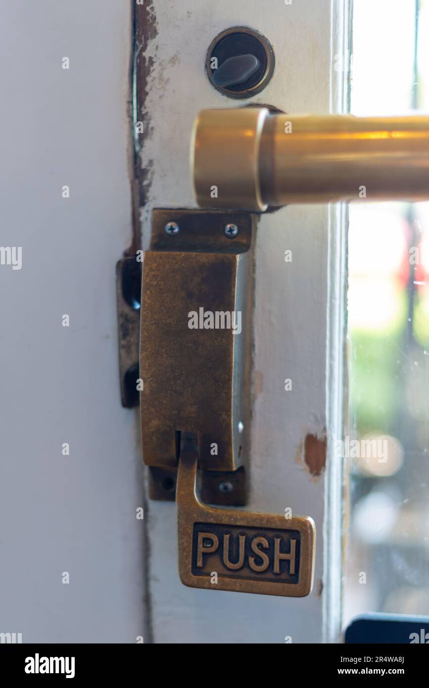 Un dispositivo di apertura della barra di spinta in ottone vintage su una porta di legno bianco con una grande finestra di vetro. La maniglia usurata ha la parola "premere" su una manopola di metallo usurata. Foto Stock