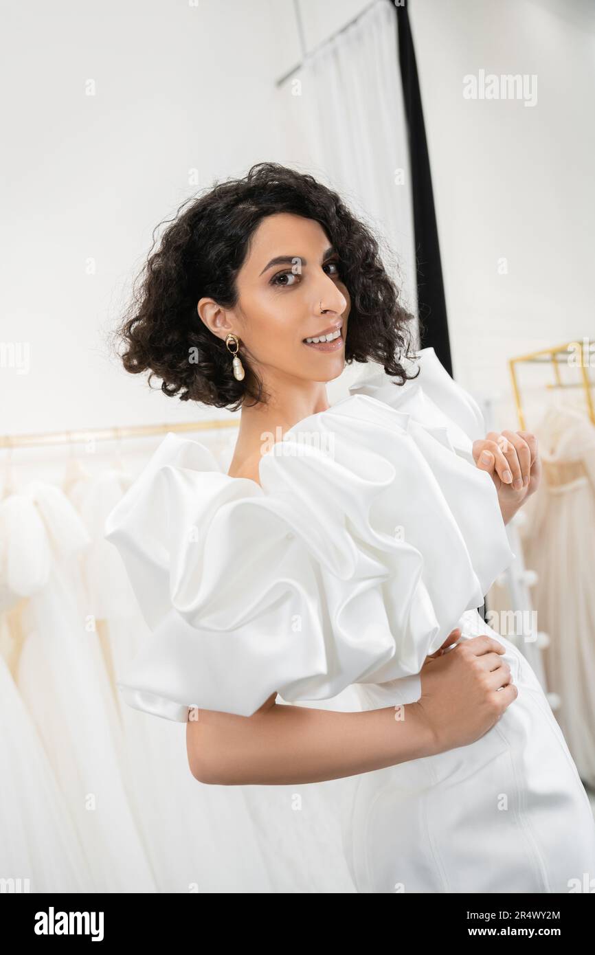 felice sposa mediorientale con brunette e capelli ondulati posati con mani sui fianchi in abito da sposa alla moda con maniche di puff e volant in buti nuziali Foto Stock