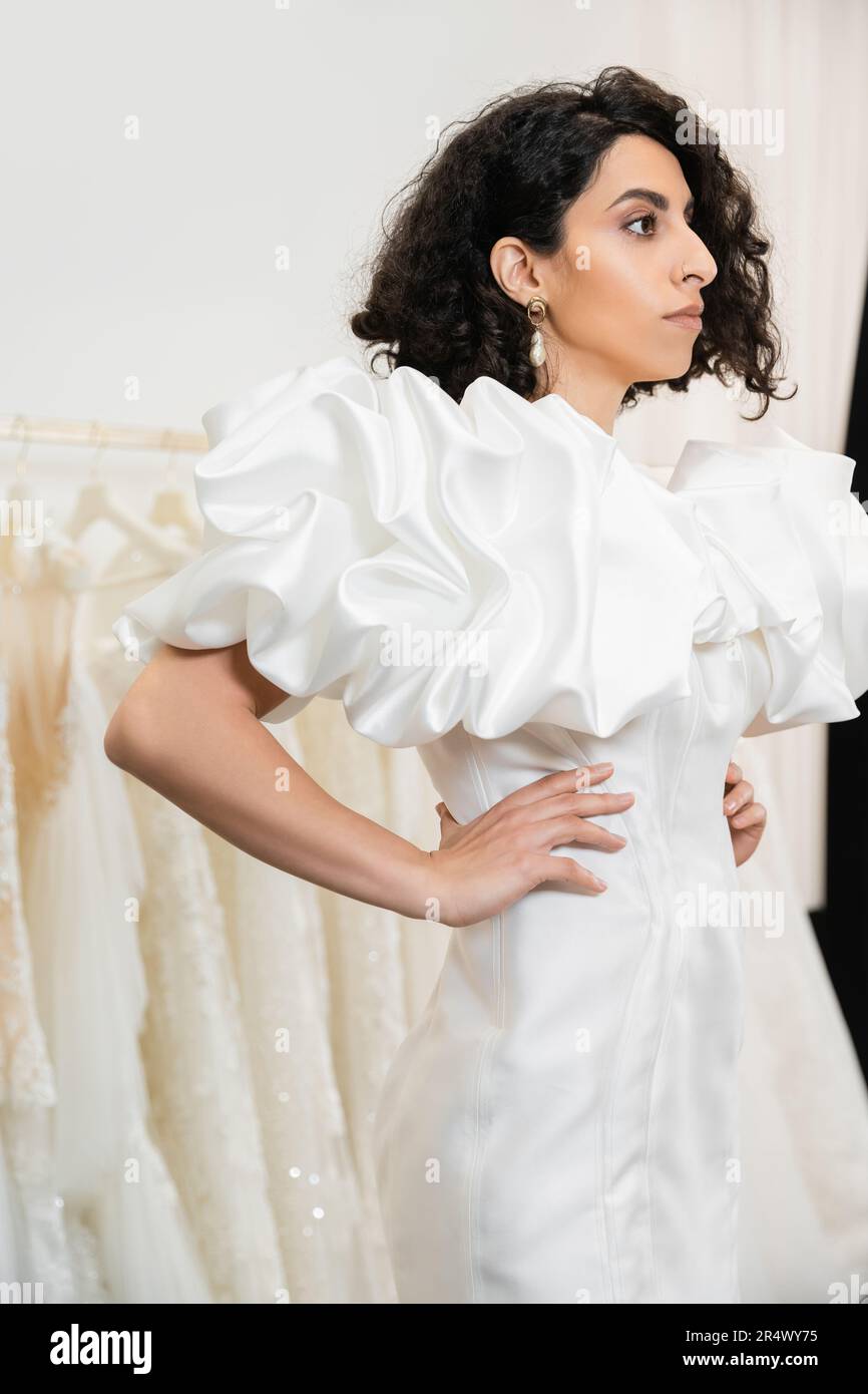 bruna sposa mediorientale con brunette e capelli ondulati che si posano con le mani sui fianchi in abito da sposa alla moda con maniche in puff e volant in bridal sa Foto Stock
