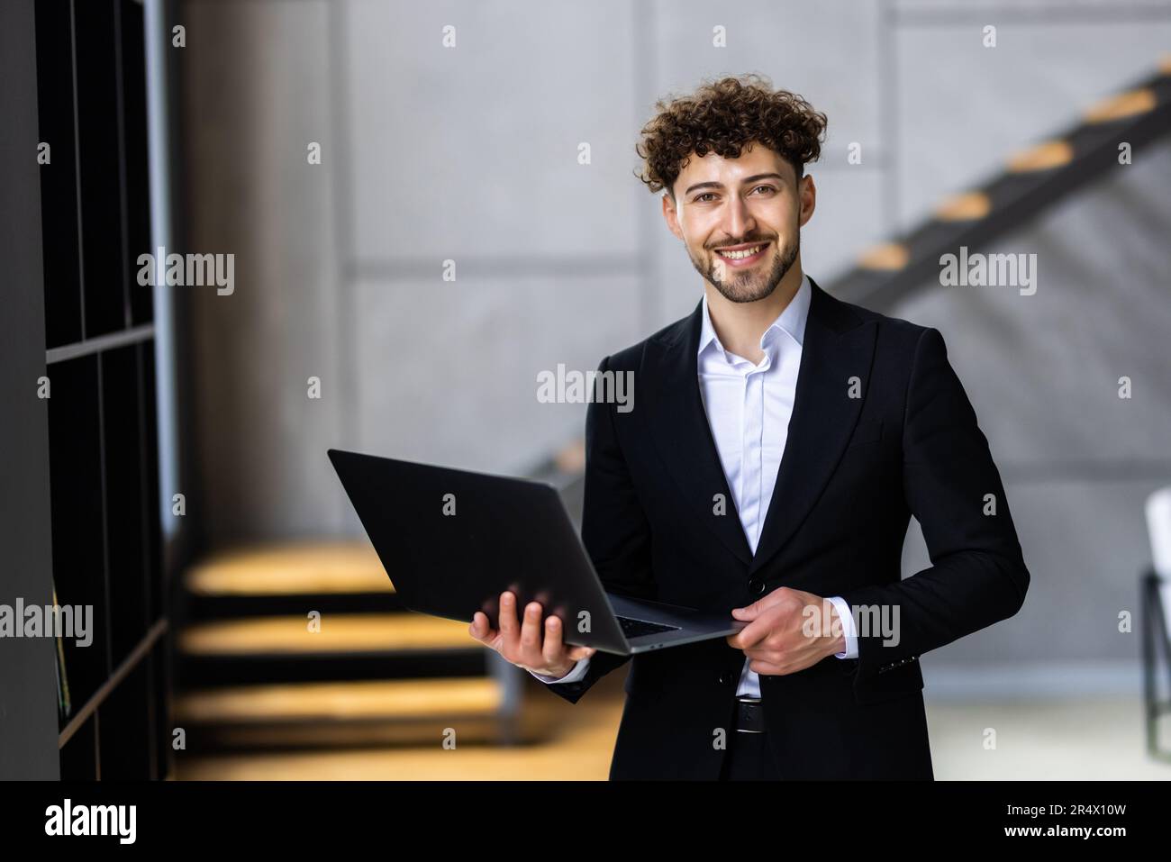 Giovane uomo d'affari moderno che indossa un blazer blu e una camicia casual, in piedi con un laptop aperto in mano Foto Stock