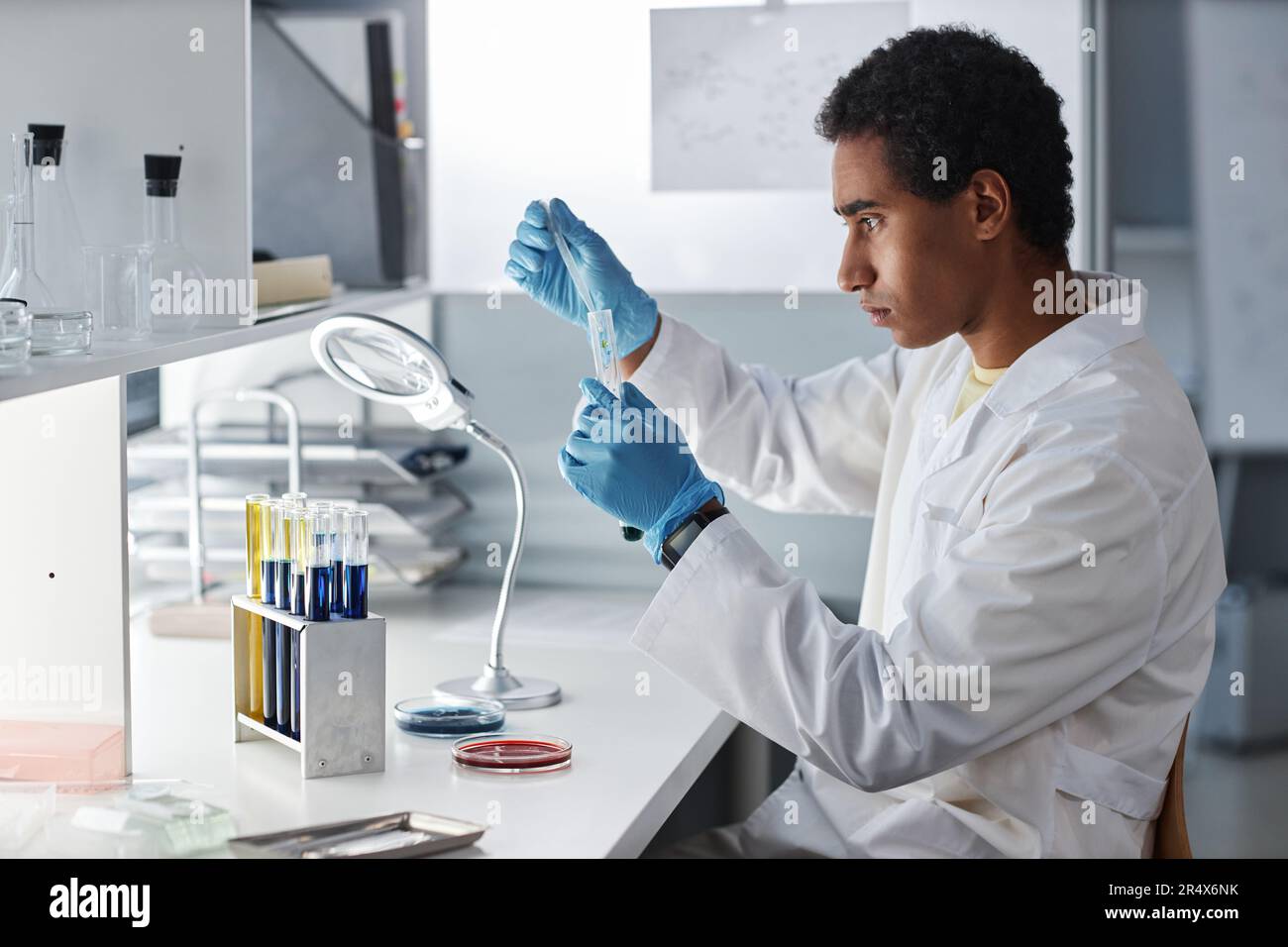 Vista laterale ritratto di giovane scienziato etnico che fa prove ed esperimenti presso la stazione di lavoro in moderno laboratorio, spazio copia Foto Stock
