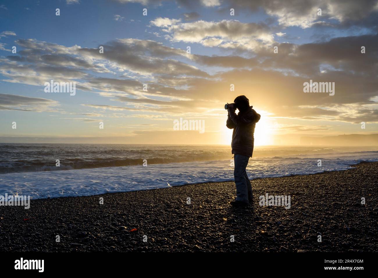 Silhouette di un fotografo in piedi sulla spiaggia che scatta una foto della vista dell'Oceano Atlantico settentrionale al tramonto; l'Islanda Foto Stock
