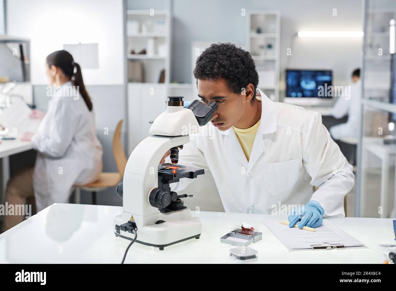 Ritratto di giovane uomo etnico come scienziato che guarda al microscopio mentre fa ricerca in laboratorio, copia spazio Foto Stock