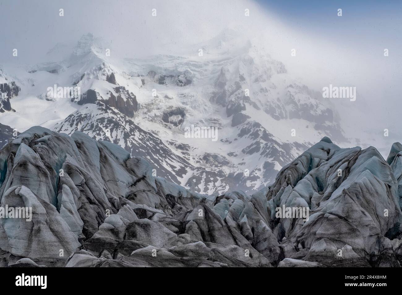 Vista panoramica delle formazioni glaciali di ghiaccio e delle montagne innevate con nuvole di nebbia che avvolgono le cime sullo sfondo; Islanda Foto Stock