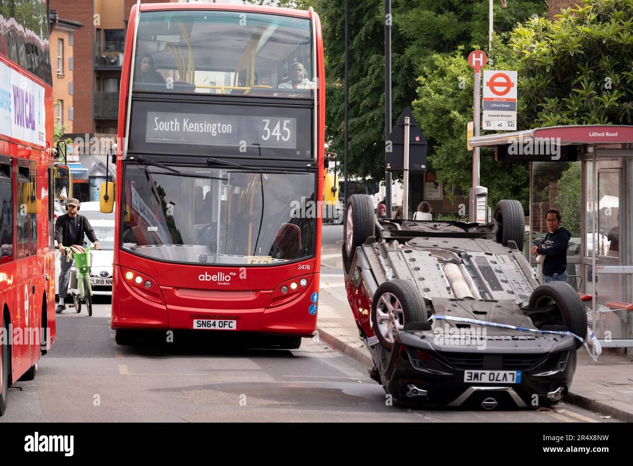 Un autobus si avvicina alle conseguenze di un incidente automobilistico in cui una berlina Nissan ribaltata rimane sul lato della strada tra Brixton e Camberwell su Coldharbour Lane, il 30th maggio 2023, a Londra, Inghilterra. Foto Stock