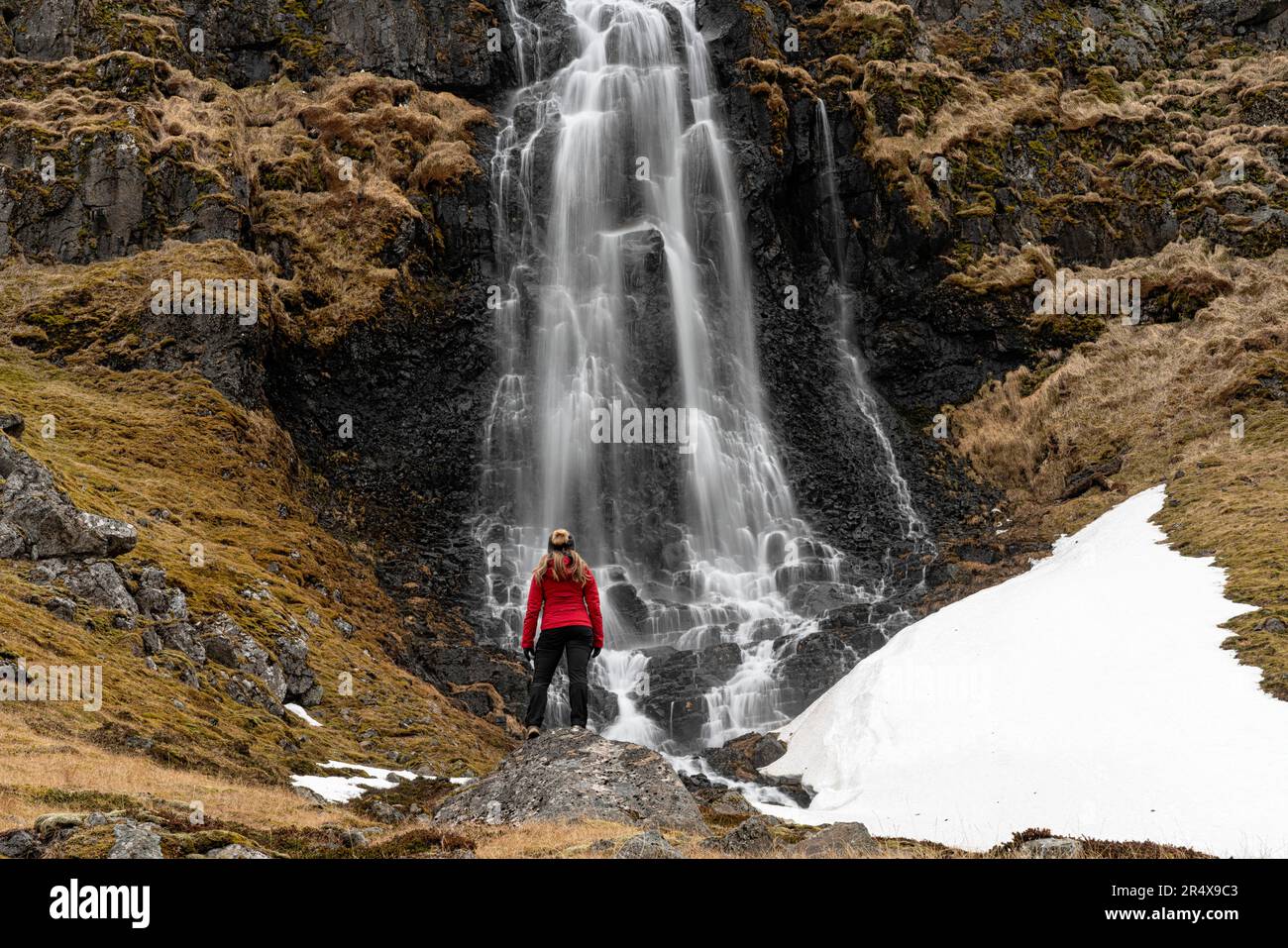 Vista da dietro di una donna in piedi di fronte a una cascata lungo la costa di Strandir vicino alla città di Djupavik, a nord-ovest dell'Islanda Foto Stock