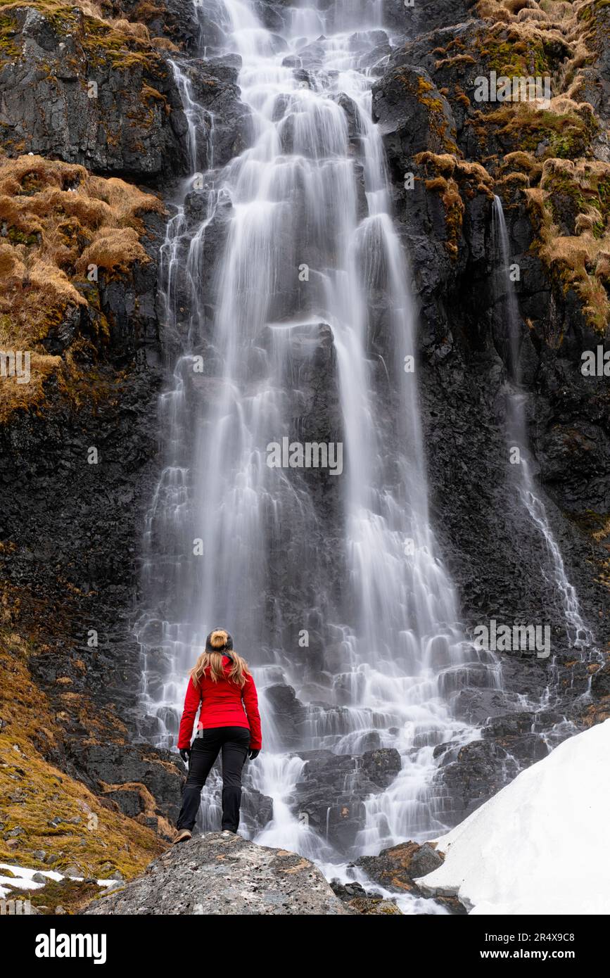 Vista da dietro di una donna in piedi di fronte a una cascata lungo la costa di Strandir vicino alla città di Djupavik, a nord-ovest dell'Islanda Foto Stock