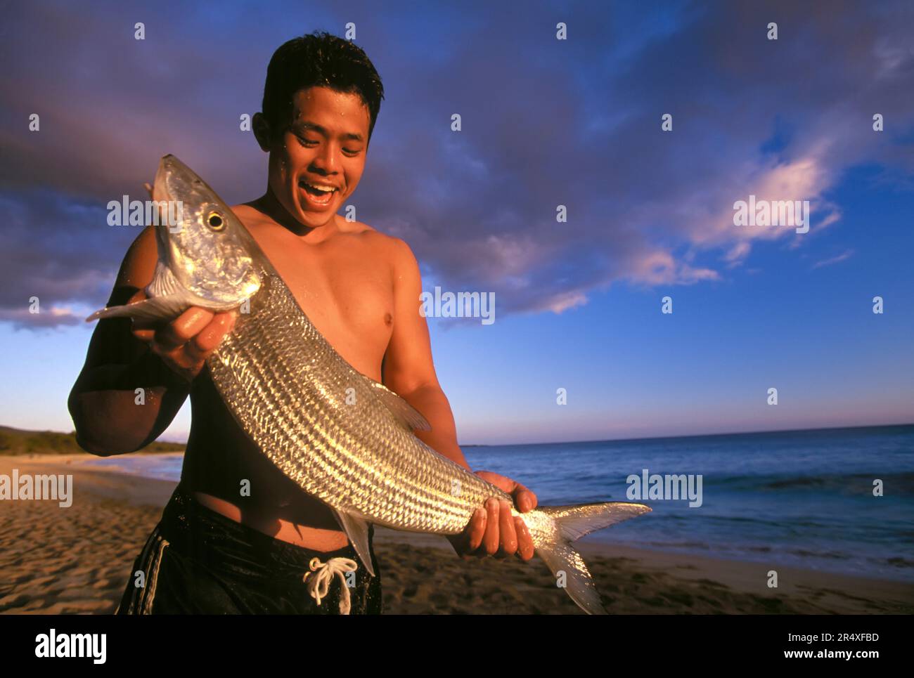 Giovane che tiene in mano un pesce rosso appena pescato su una spiaggia al crepuscolo; Maui, Hawaii, Stati Uniti d'America Foto Stock