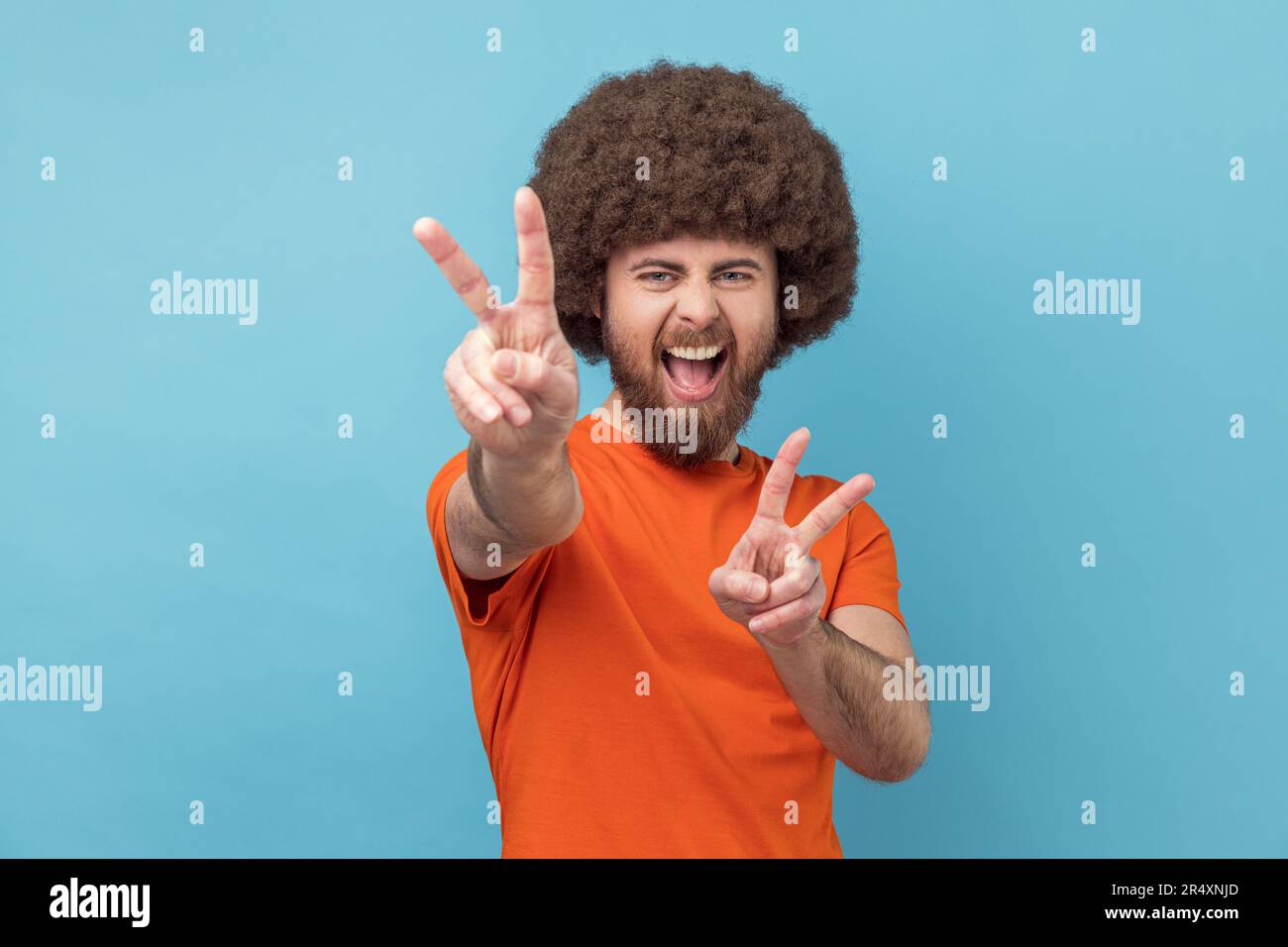 Ritratto di eccitato allegro bell'uomo con acconciatura Afro indossare T-shirt arancione mostrando segno di pace, v gesto e urlare felicemente. Studio in interni isolato su sfondo blu. Foto Stock