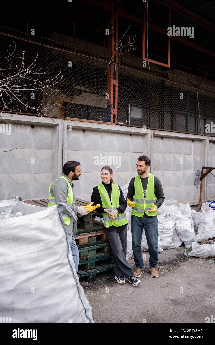 Sorridente lavoratore indiano in gilet ad alta visibilità e guanti che parlano con colleghi allegri, mentre in piedi vicino a sacchi con rifiuti in smaltimento di rifiuti all'aperto Foto Stock