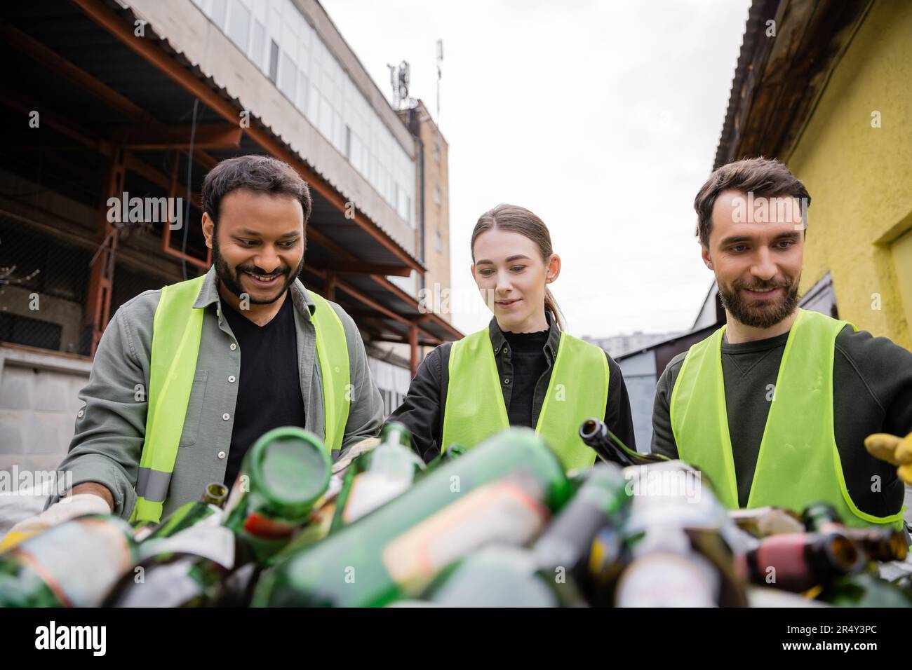Smistatori interrazziali positivi in giubbotti protettivi che lavorano con rifiuti in vetro sfocato in stazione di smaltimento rifiuti all'aperto, raccolta rifiuti e riciclaggio con Foto Stock