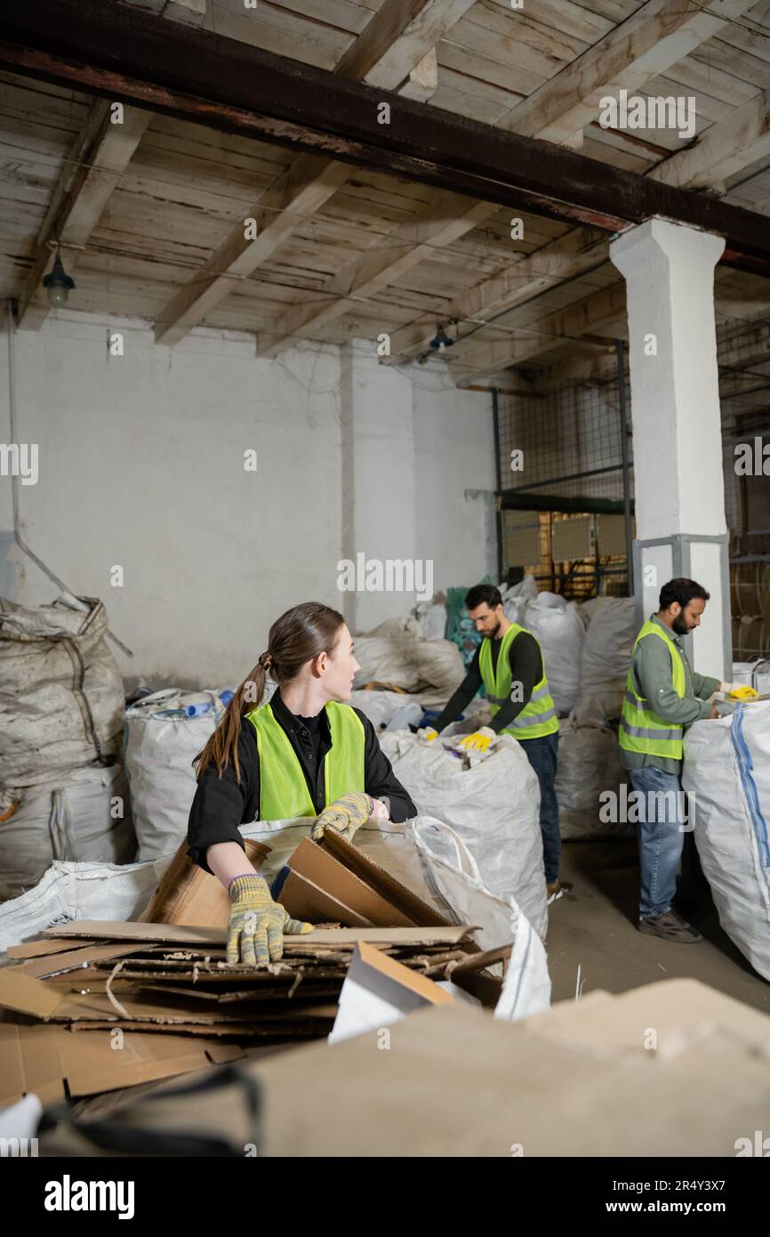 Giovane lavoratrice in guanti protettivi e giubbotto guardando i colleghi multietnici mentre mette il cartone nel sacco nella stazione di smaltimento rifiuti, rifiuti Foto Stock