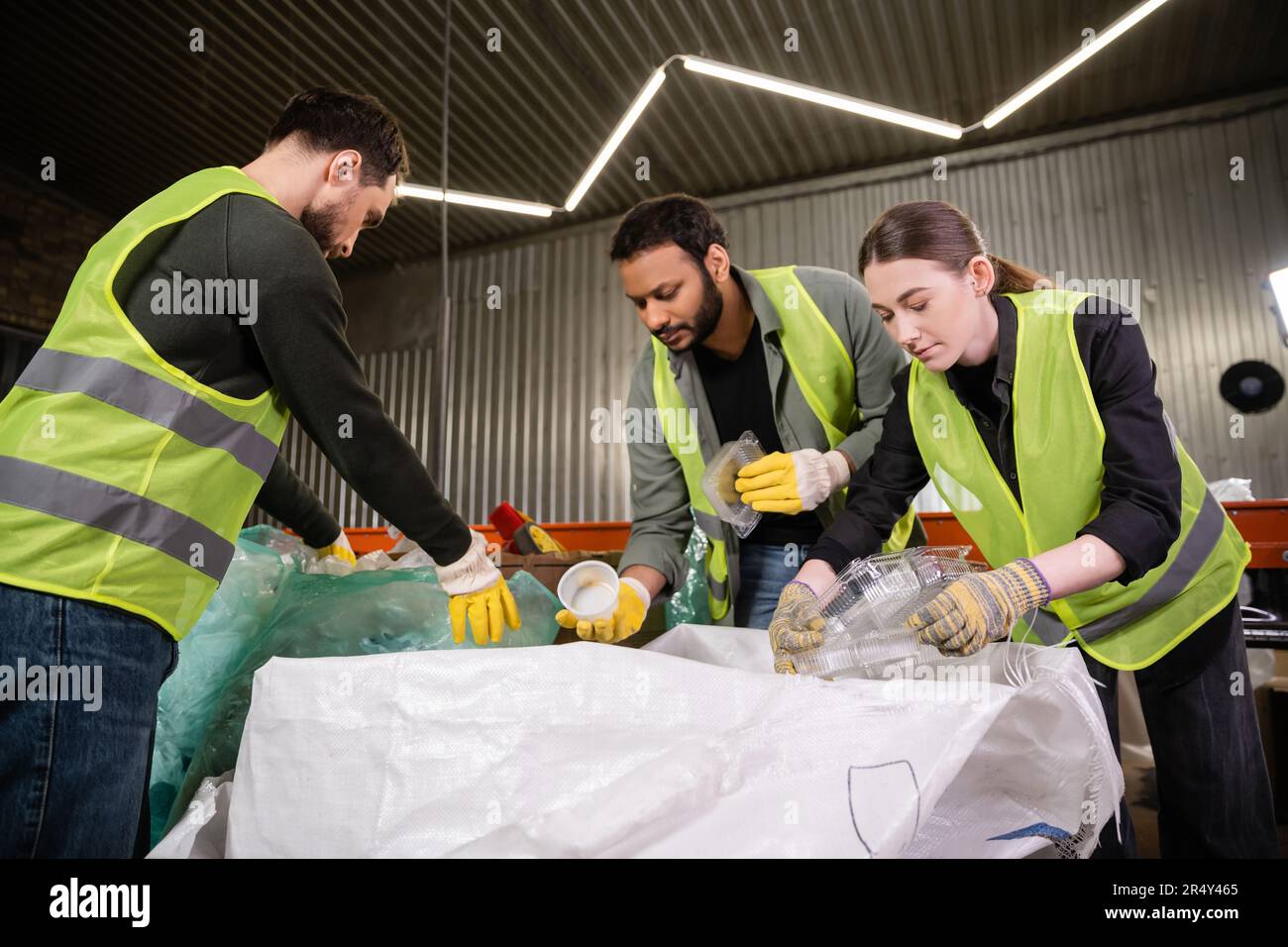 Giovane lavoratore in giubbotto riflettente e guanti che tengono contenitori di plastica vicino al sacco mentre smistamento rifiuti con colleghi multietnici in stat di smaltimento rifiuti Foto Stock