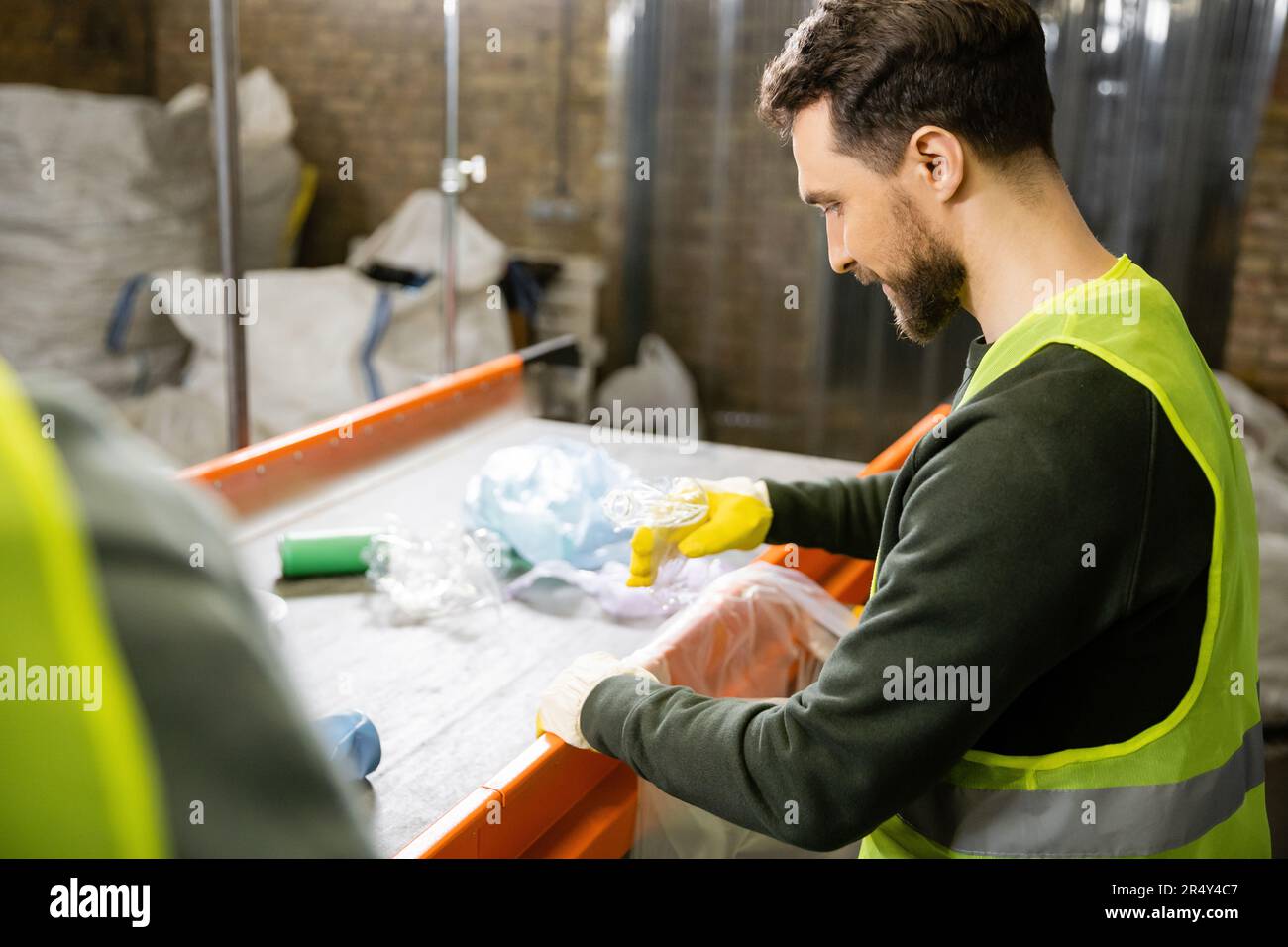 Vista laterale del lavoratore in guanti protettivi e giubbotto di smistamento rifiuti vicino al trasportatore mentre si trova in una stazione di smaltimento rifiuti sfocati sullo sfondo, garbag Foto Stock