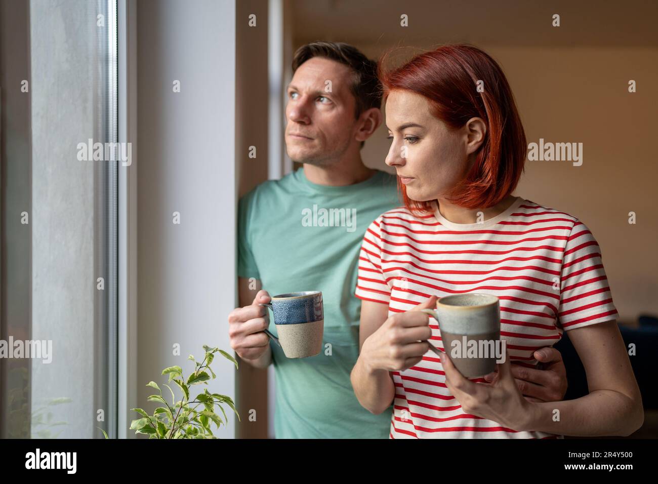 Preoccupazione seria giovane coppia della famiglia in piedi insieme dalla finestra a casa che beve il caffè di mattina Foto Stock