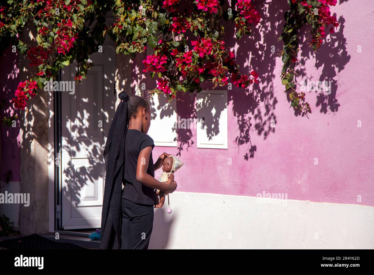 Bambino che tiene in mano una bambola davanti a un muro rosa con fiori di Bougainvillea Foto Stock
