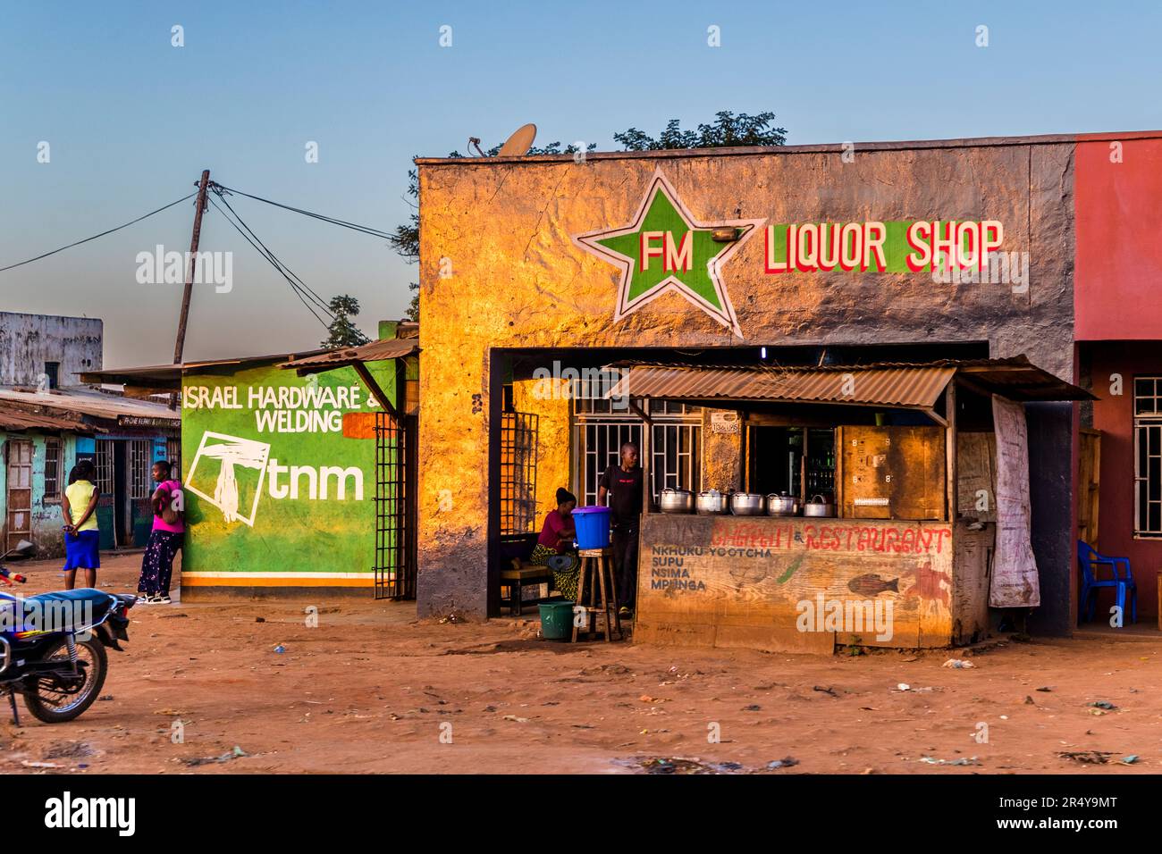 Un "FM Liquor Shop" e una bancarella alimentare adiacente con un menu dipinto a mano – Nkhuku Yotcha (pollo grigliato), Supu (zuppa), Nsima (porridge di mais, cibo base del Malawi) e Mpunga (riso) – si trovano lungo una strada a Lilongwe, Malawi, al crepuscolo. Un'insegna vicina pubblicizza "Israel Hardware & Welding". Foto Stock