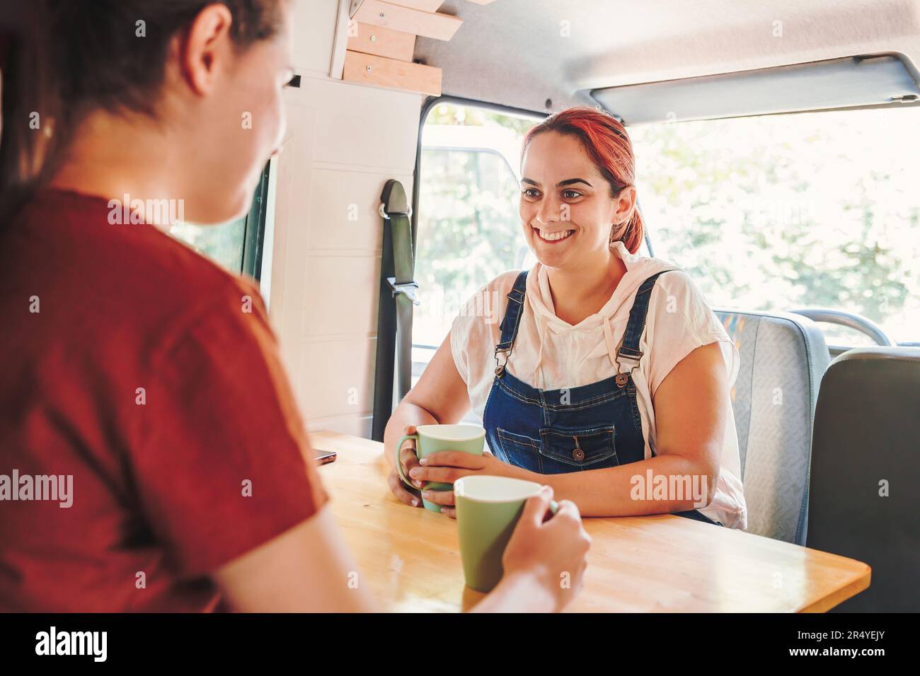 Due giovani donne bevono caffè e si sono fatte un bel po' di tempo in un camper fatto a mano. Viaggio in pulmino e avventura estiva all'aperto. Stile di vita Nomad Foto Stock