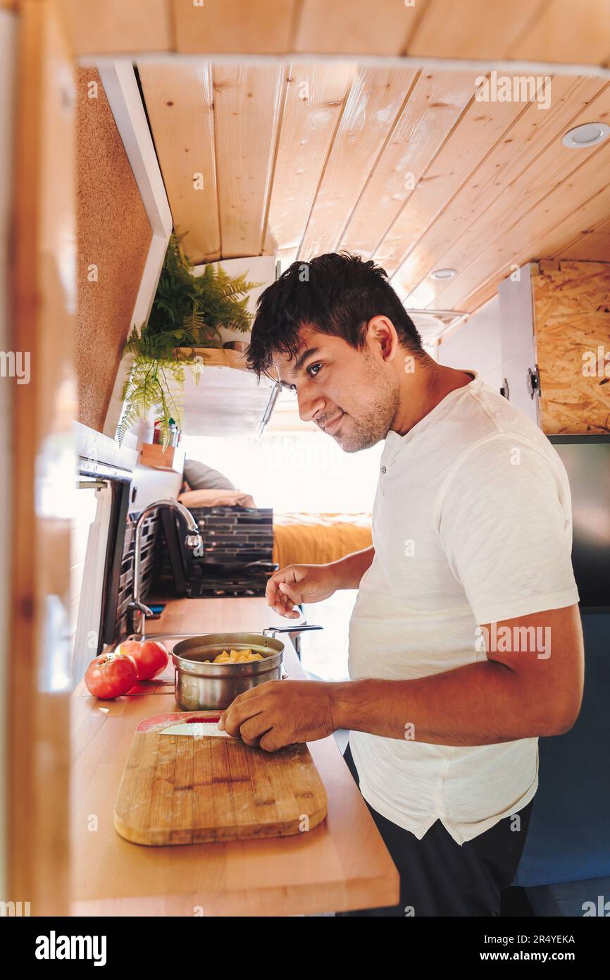 Un uomo ispanico che cucina il cibo per il pasto nella cucina del suo camper. Viaggio in pulmino e avventura estiva all'aperto. Concetto di stile di vita Nomad Foto Stock