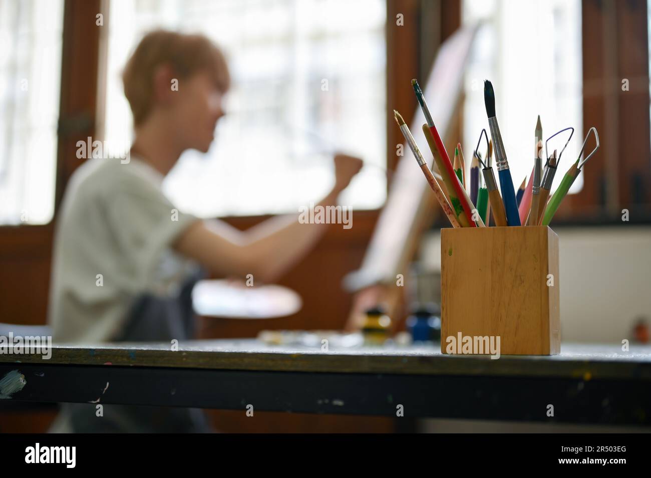 Immagine ravvicinata di un supporto a matita con varie matite e strumenti di pittura su un tavolo su uno sfondo sfocato di un uomo dipinto su una tela. selettivo Foto Stock