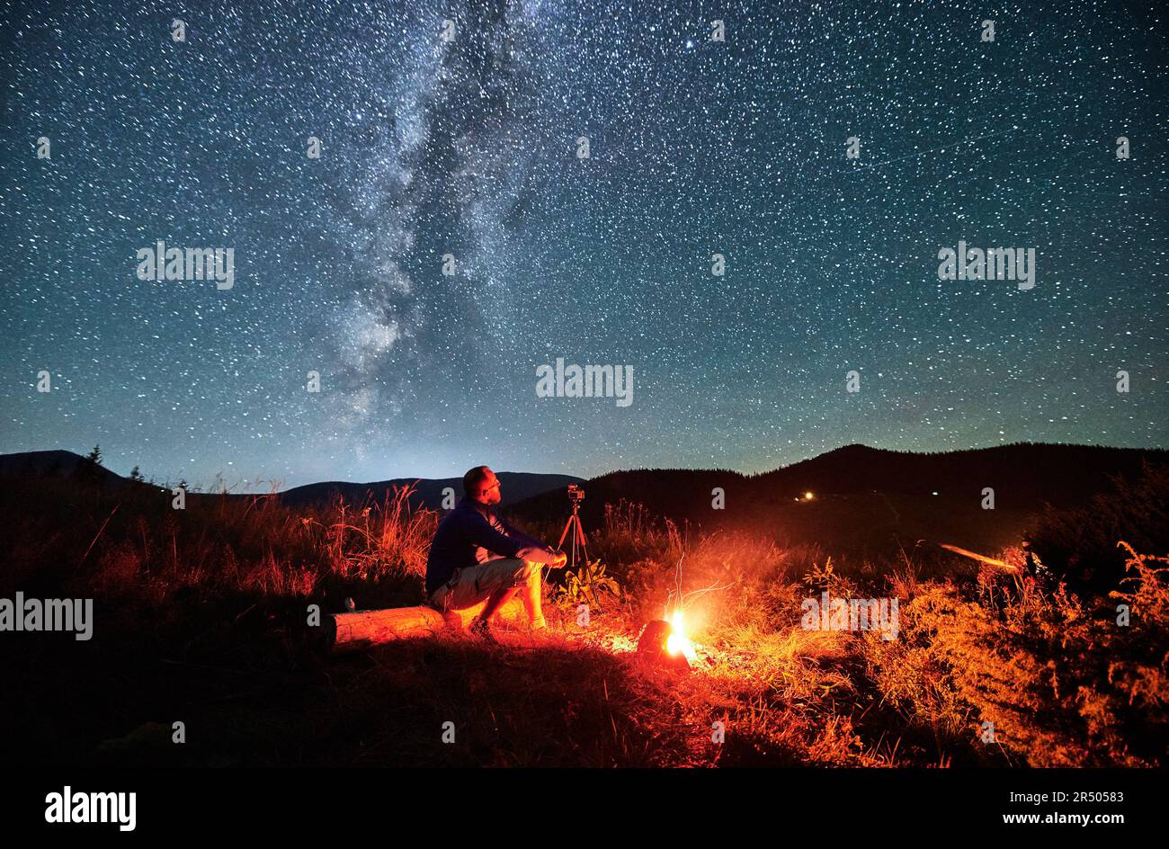 Uomo che scatta foto di cielo stellato con la Via Lattea. Fotografo maschile seduto sul log e fotocamera di messa a fuoco. Uomo con macchina fotografica seduto accanto al fuoco del campo in montagna. Foto Stock
