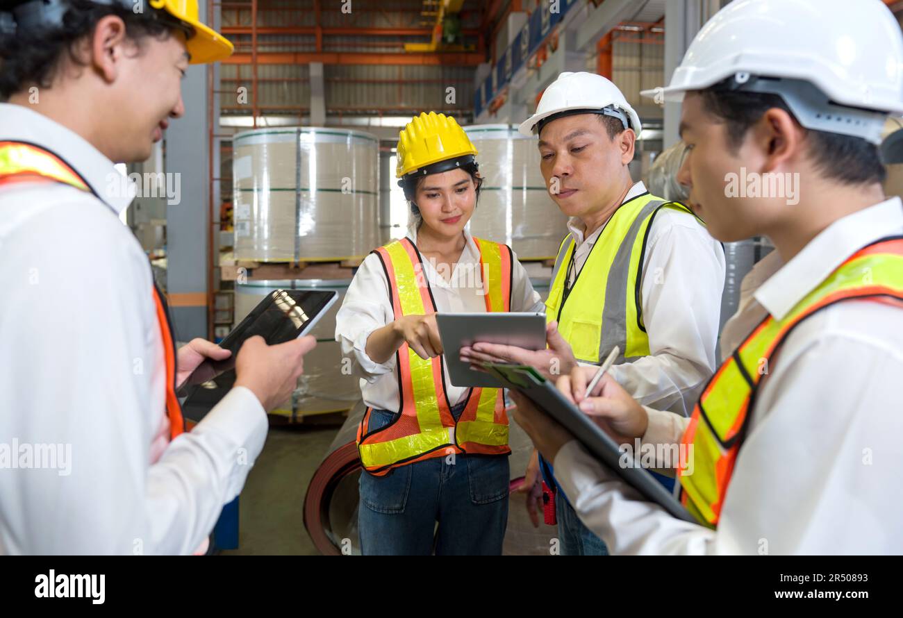 Il Senior Engineer Manager forma i nuovi dipendenti all'interno della fabbrica di lamiere. Tutti indossano giubbotto di sicurezza e elmetto. La pila di rulli in lamiera di acciaio si trova in Foto Stock