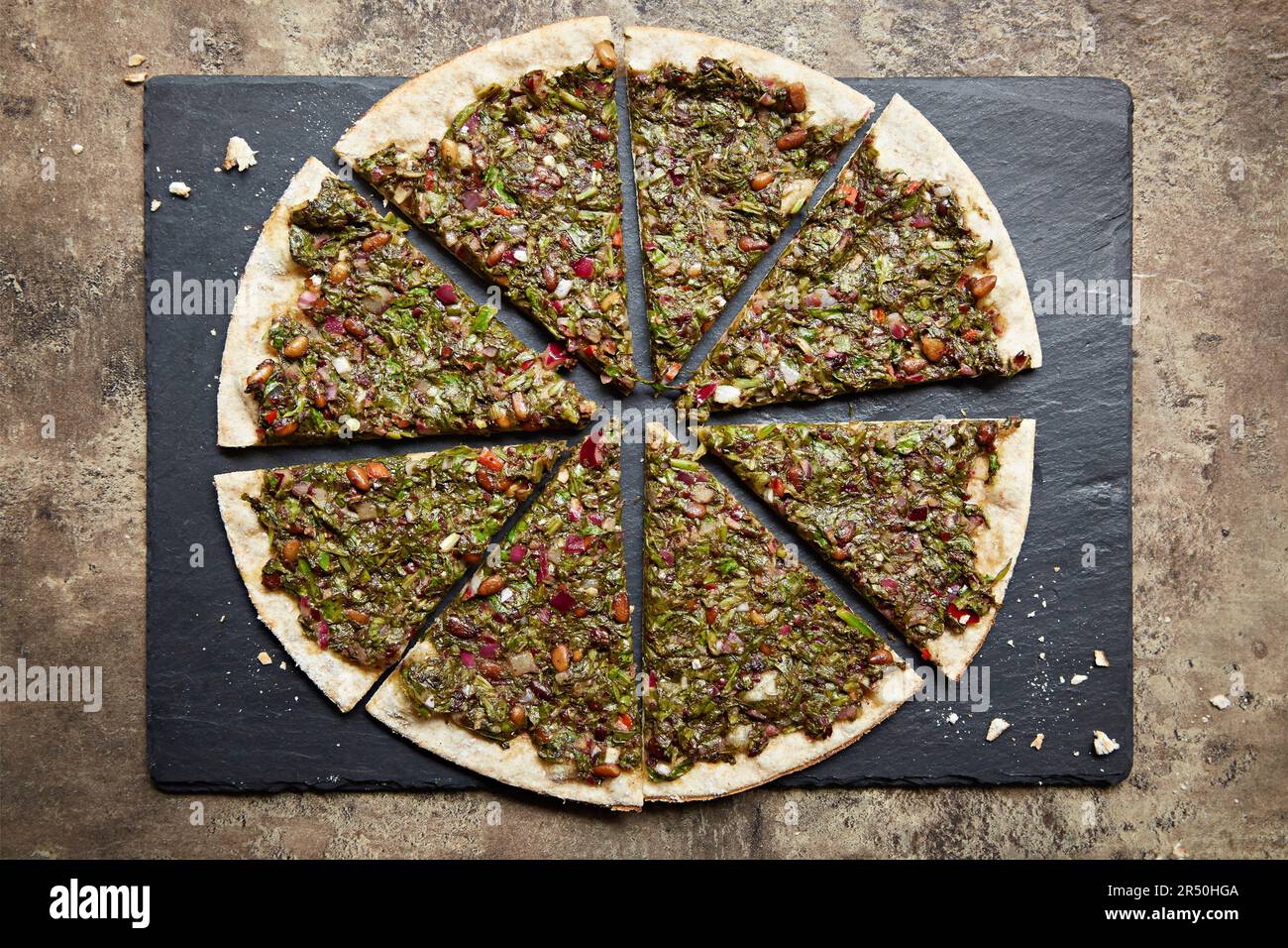 Una pizza sormontata con verdure a foglia, pistacchi, pinoli e melograno, affettata su un piatto di ardesia Foto Stock