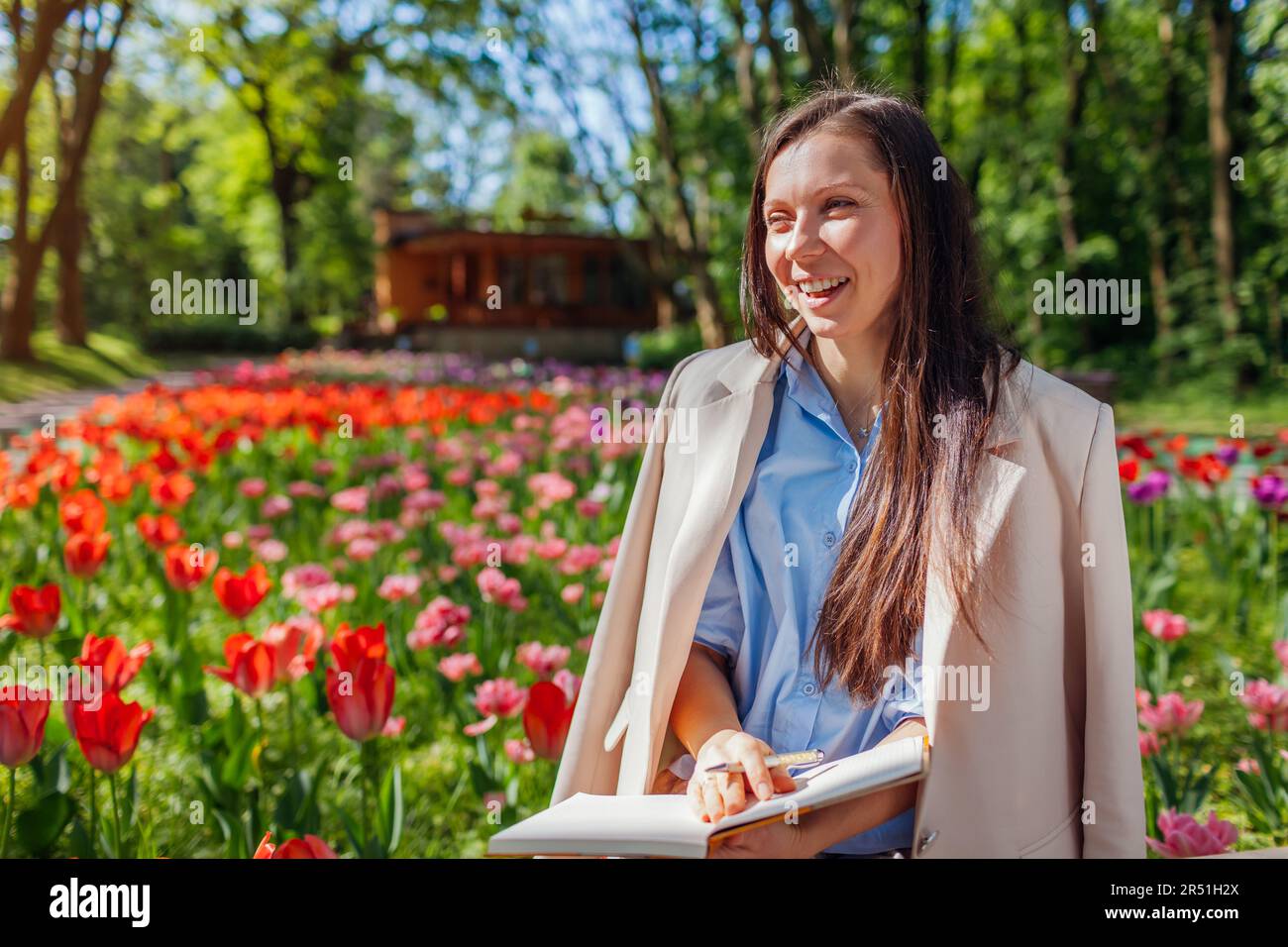 Giovane donna d'affari caucasica che ride tenendo notebook in primavera parco. Gli insegnanti hanno una pausa all'aperto rilassandosi nel campus universitario. Spazio Foto Stock