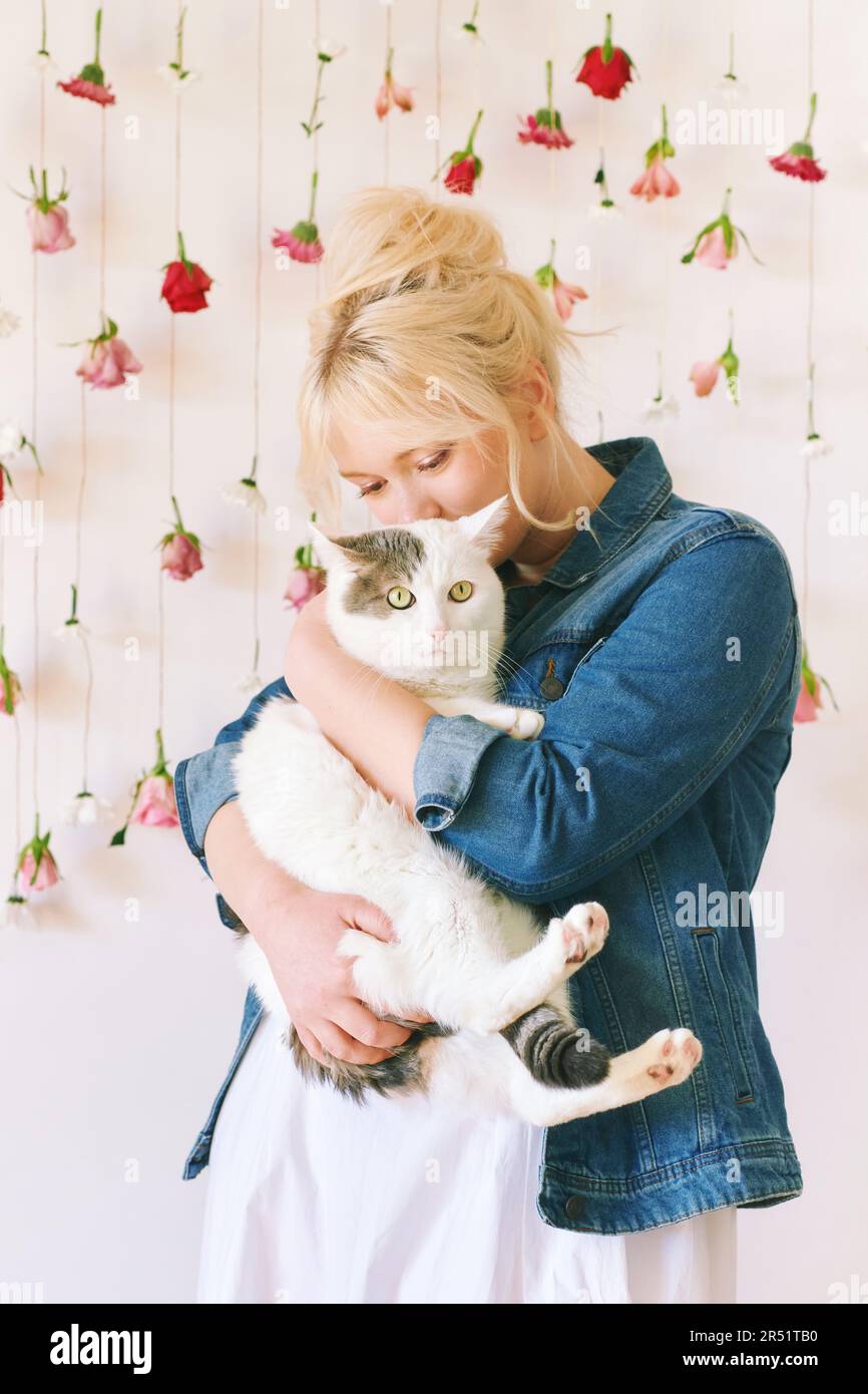 Studio ritratto di bella giovane adolescente 15 - ragazza di 16 anni che indossa giacca denim, posa su sfondo bianco con fiori appesi, tenendo carino gatto, Foto Stock