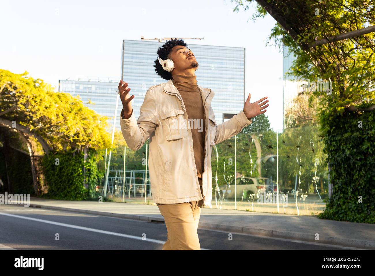 Felice afro americano giovane con gli occhi chiusi ascoltando musica con cuffie e braccia aperte mentre cammina per la strada contro gli alberi Foto Stock