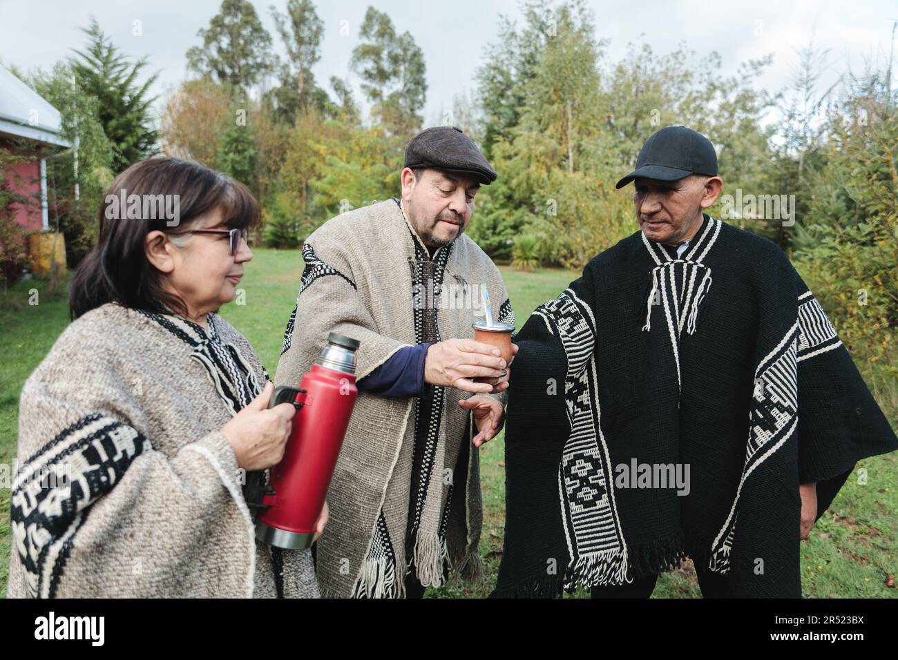 Gruppo di amici in abiti caldi in piedi vicino a cespugli verdi e versare una bevanda calda da thermos in tazza, mentre si passa il tempo insieme nella giornata di sole Foto Stock