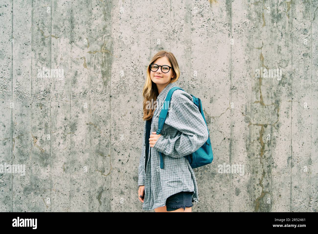 Ritratto all'aperto della giovane ragazza adolescente che indossa occhiali e zaino, posato su sfondo grigio parete Foto Stock
