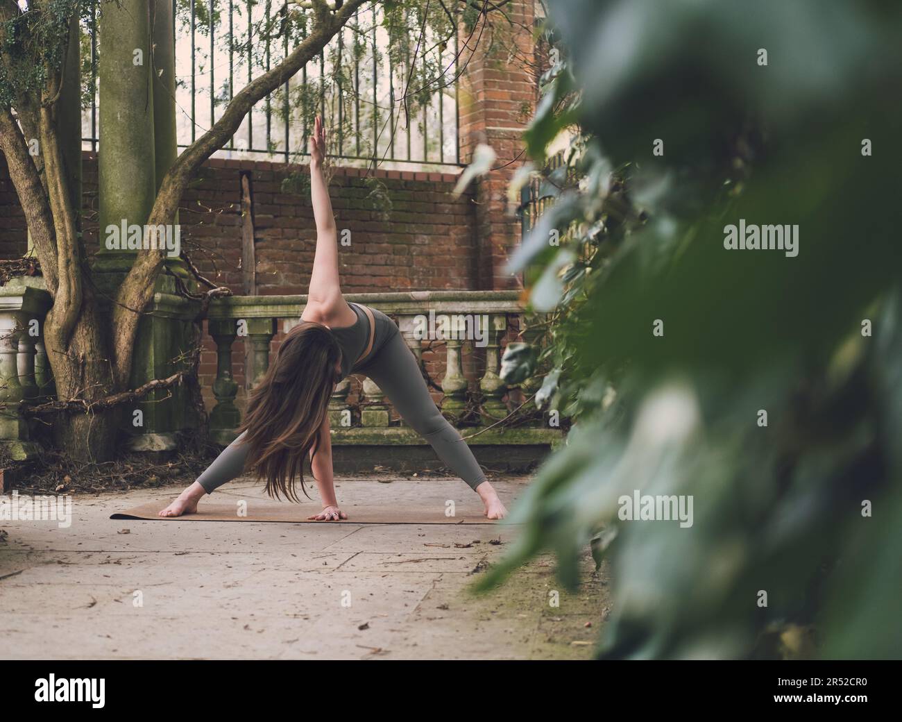 Foto artistica di una donna irriconoscibile insegnante di yoga in Revolved o Twist Wide Legged Forward Bend posa in una terrazza di uno storico giardino paesaggistico Foto Stock