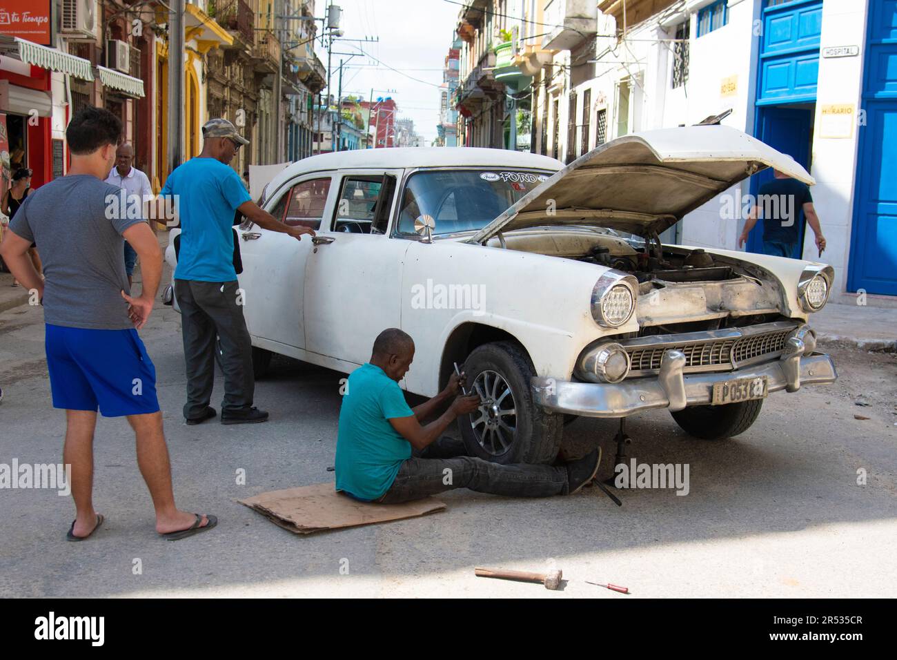 L'uomo cubano fissa il pneumatico sulla sua auto a l'Avana, Cuba, mentre altre persone si fermano, guardano e parlano. Vita a Cuba. Foto Stock