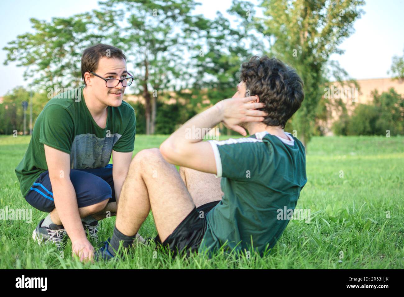 Due amici maschili che si allenano e si siedono con la t-shirt verde nel parco con l'erba nelle giornate di sole Foto Stock