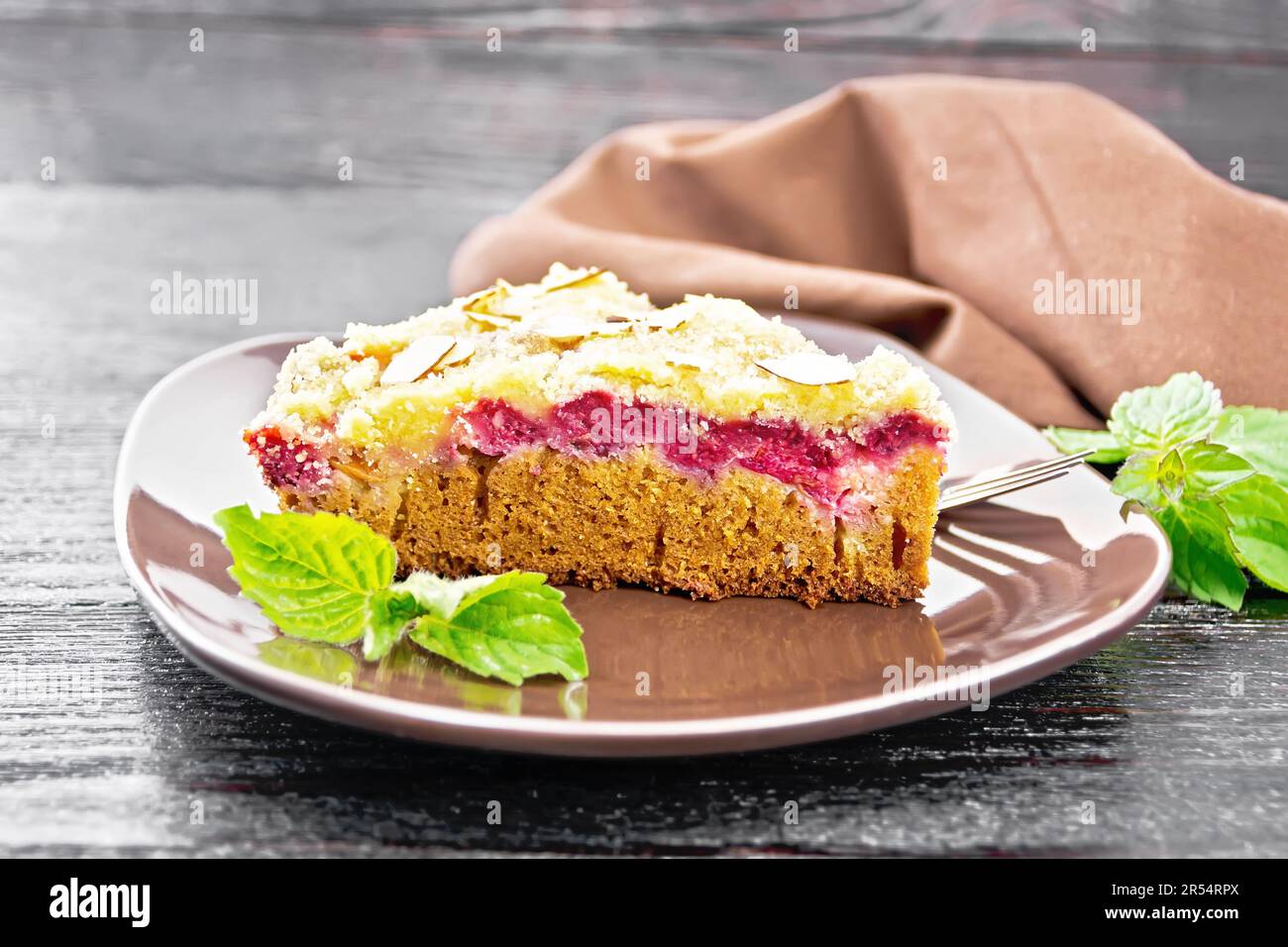 Un pezzo di dolce con lamponi, burro crumble e fiocchi di mandorle in un piatto, menta e asciugamano su fondo di legno scuro Foto Stock
