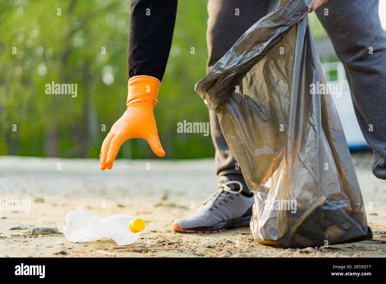 Primo piano della mano maschile in guanti di gomma con sacco per rifiuti che raccoglie una bottiglia di plastica sulla spiaggia. Concetto di inquinamento ambientale. Giornata della Terra. Foto Stock
