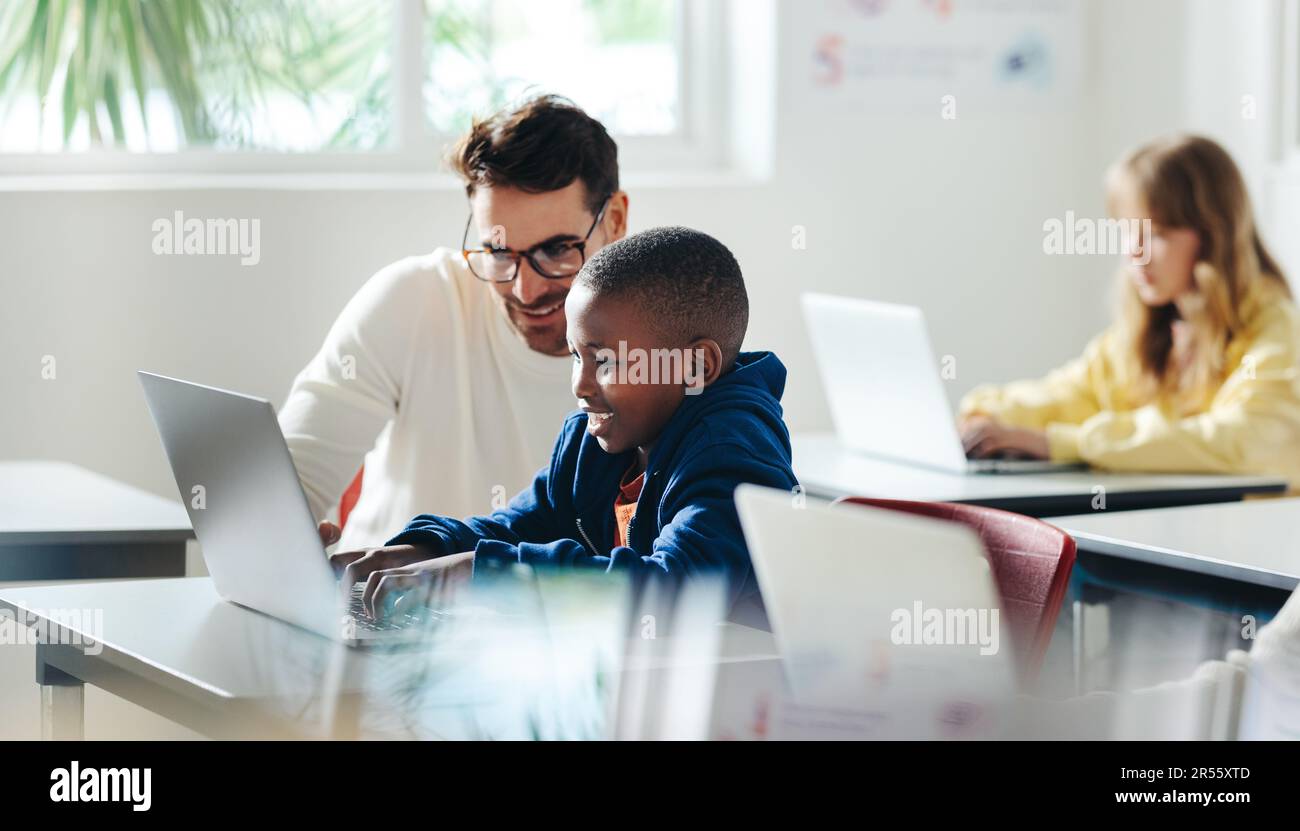 Un insegnante di sesso maschile aiuta un giovane ragazzo con l'apprendimento basato sul computer in un ambiente scolastico. Tutor infantile che fornisce una lezione in una scuola elementare, con un foc Foto Stock