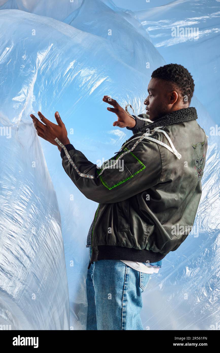 Vista laterale dell'elegante uomo afroamericano in giacca con righe a LED e jeans strappati che toccano il cellophane lucido su sfondo blu, outfit urbano e. Foto Stock
