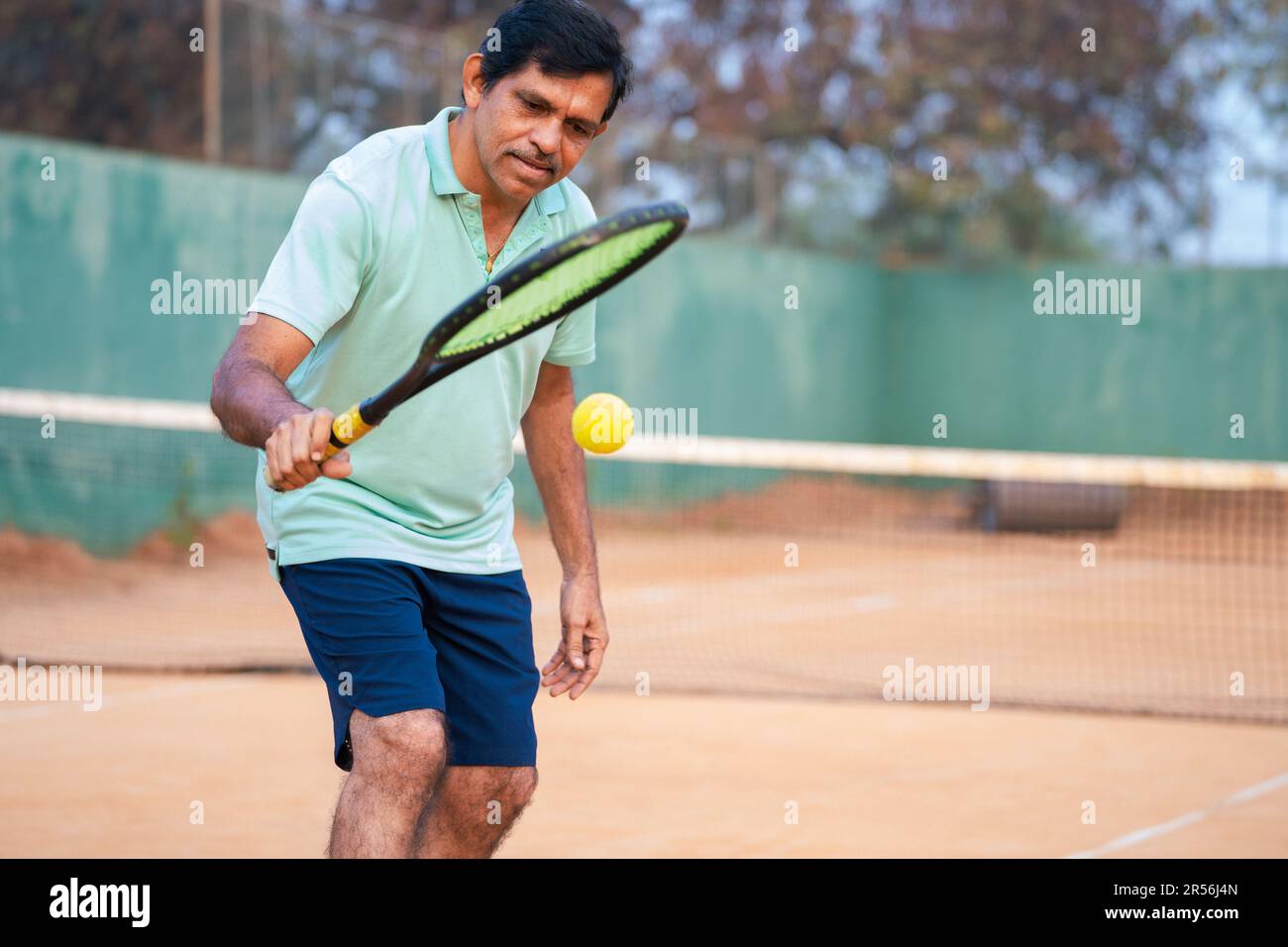 Un uomo anziano indiano sano e attivo che pratica il tennis colpendo la palla sul groud in campo - concetto di pratica, stili di vita sani e routine mattutina. Foto Stock