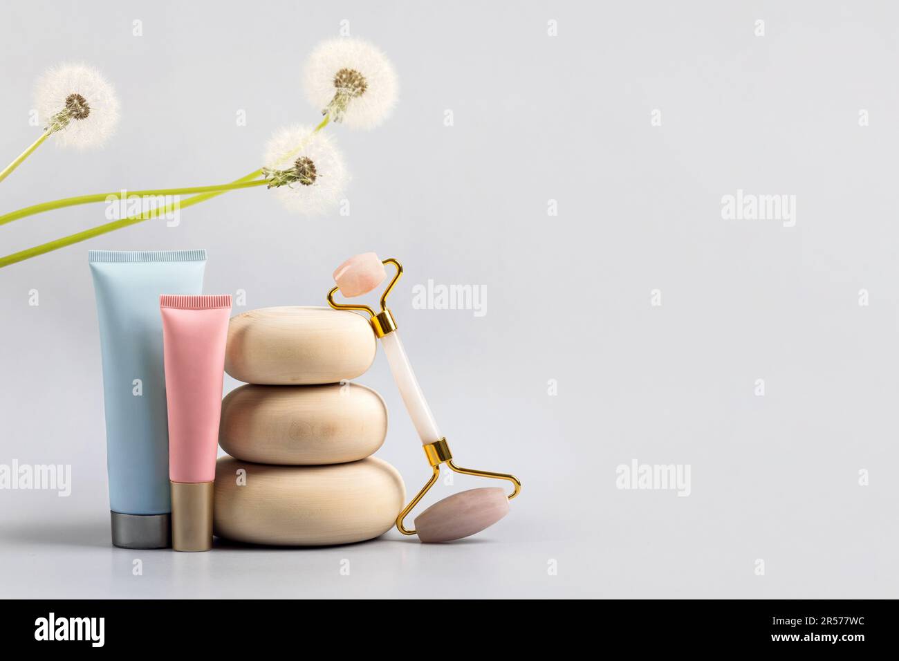 Rullo facciale in quarzo rosa e contenitore per bottiglie cosmetiche su pietre di legno con dente di leone floreale, sfondo grigio. Massaggio viso anti-età per l. Naturale Foto Stock