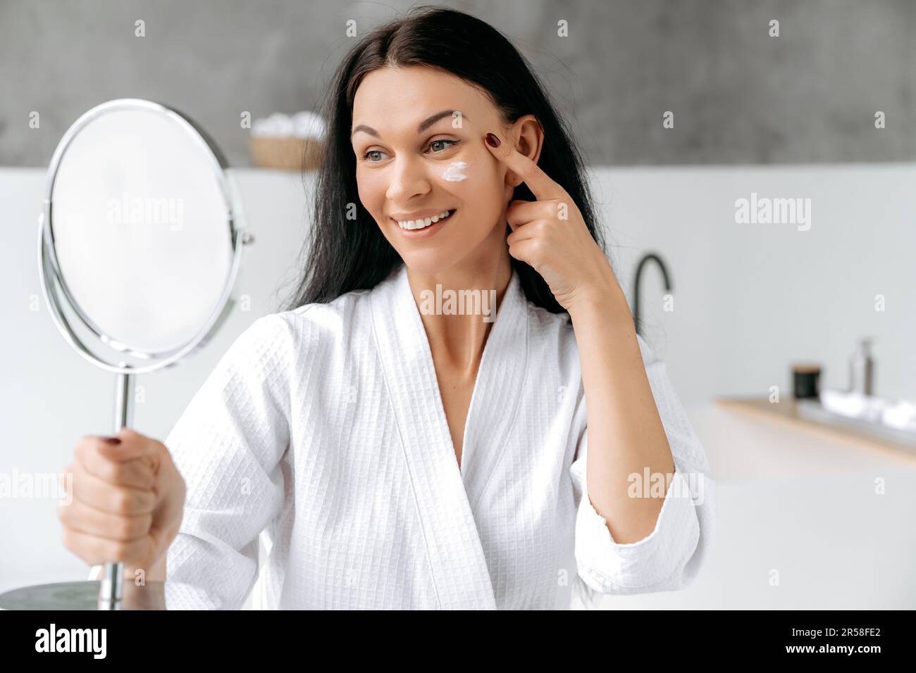 Splendida donna caucasica ben curata in un accappatoio, tiene e guarda uno specchio, applica una crema idratante anti-invecchiamento sul viso, previene l'invecchiamento della pelle e asciutto. Trattamenti giornalieri idratanti per la pelle Foto Stock