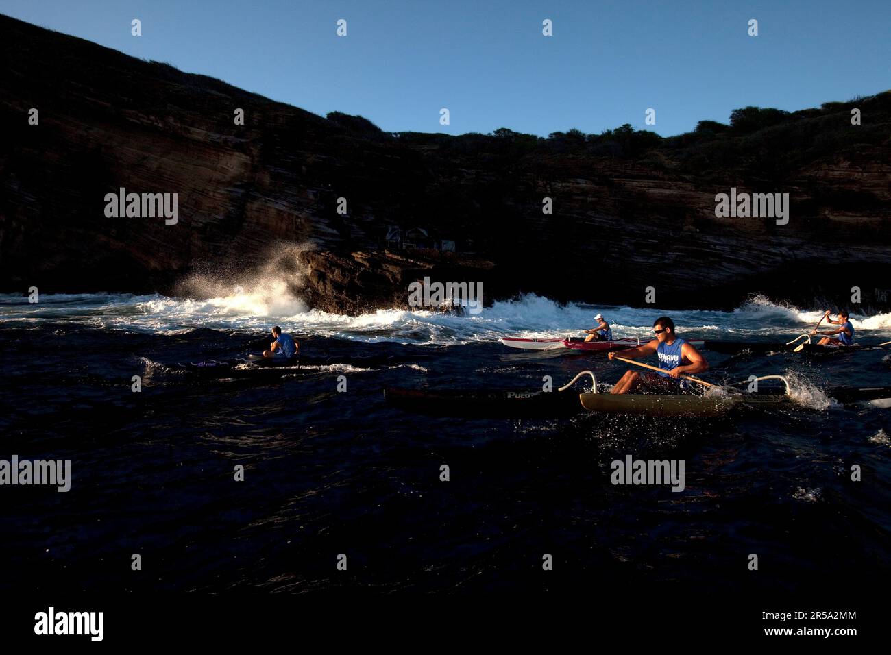 Quattro uomini in un uomo scatenano canoe che si arrampicano accanto a scogliere e onde che si infrangono. Foto Stock