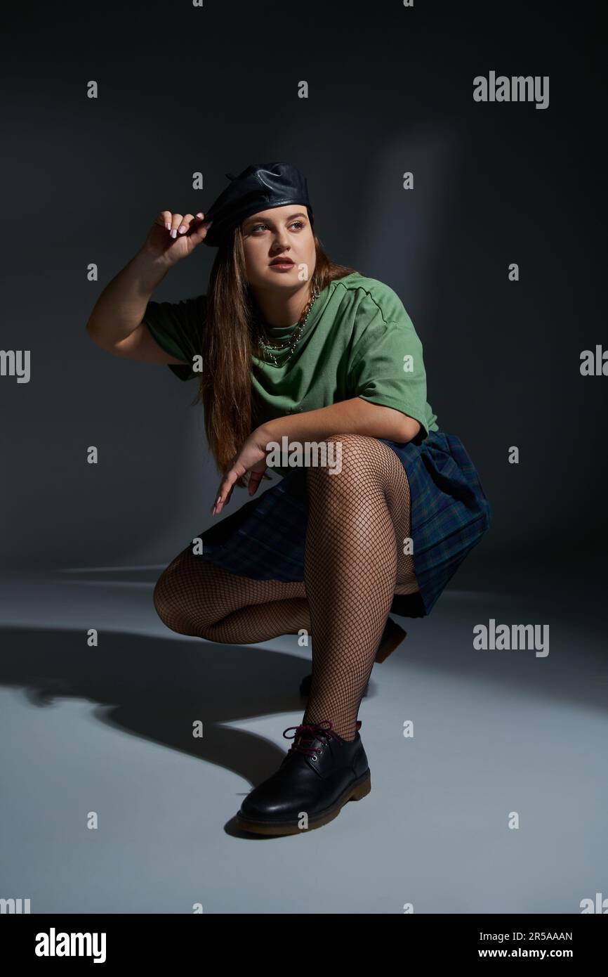 intera lunghezza di donna più taglia posa in berretto di pelle, t-shirt verde, gonna a plaid con catene, collant a rete di pesce e scarpe nere seduta e guardare awa Foto Stock
