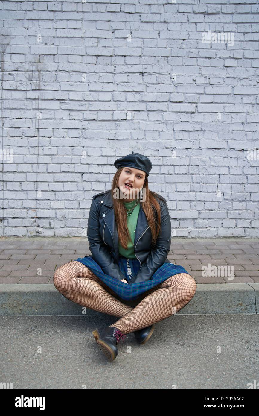 tutta la lunghezza di donna chic più misura in posa in giacca di pelle, gonna a plaid con catene, collant a rete e scarpe nere mentre seduto vicino a parete di mattoni Foto Stock