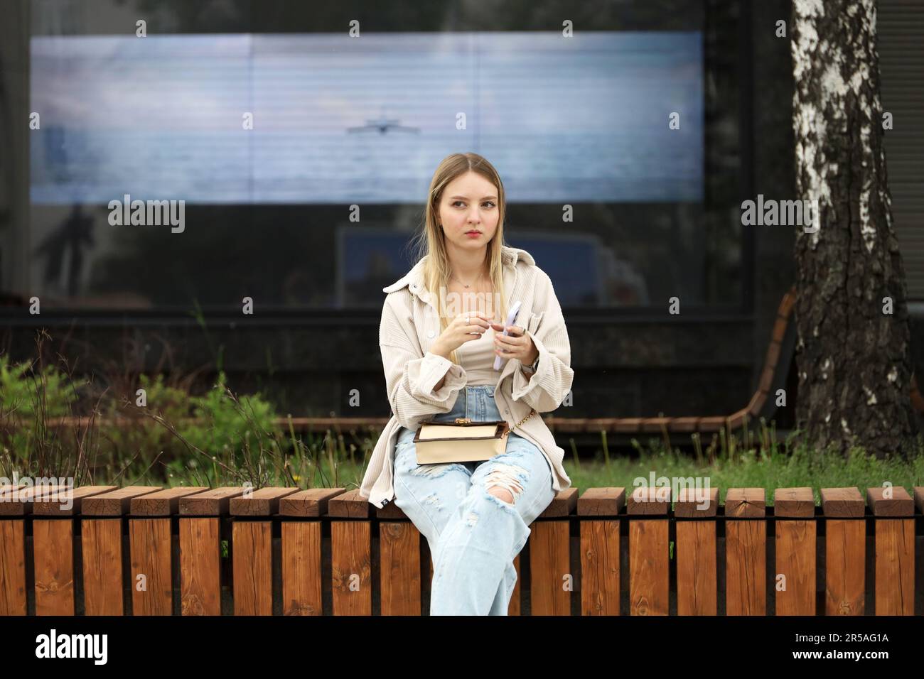 Ragazza bionda in jeans strappati con smartphone seduto su una panca di legno sulla strada cittadina Foto Stock
