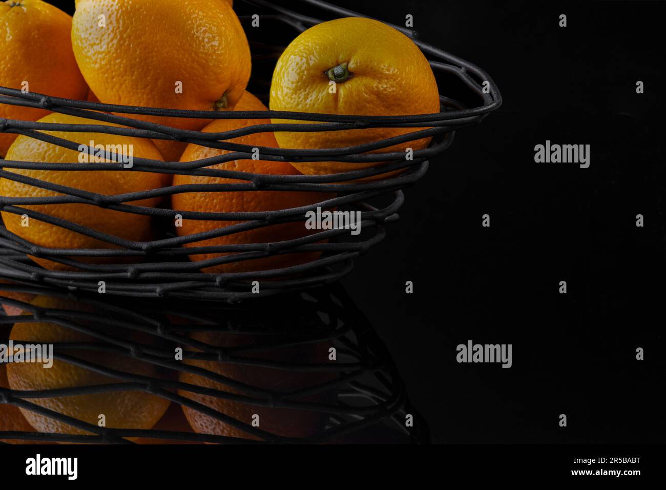 un cesto di filo nero con arance fresche su sfondo nero a specchio. Foto Stock