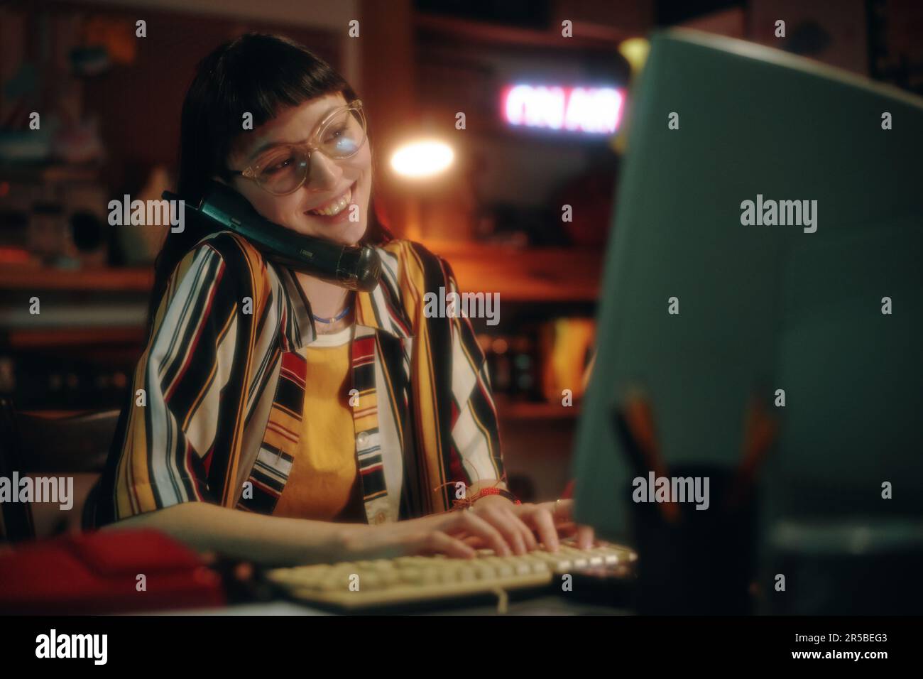 Giovane donna sorridente che ha parlato al telefono mentre lavora al computer al tavolo in officina Foto Stock